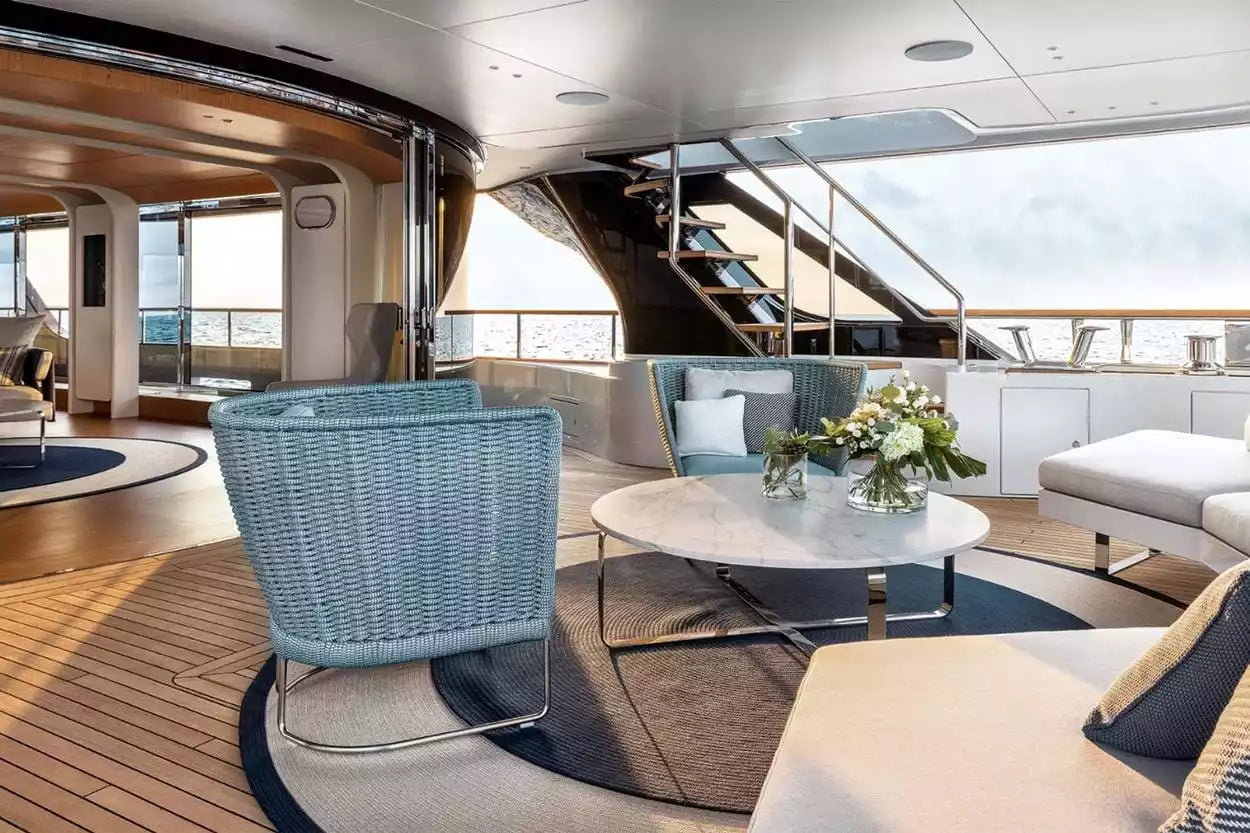 A luxury yacht or resort lounge interior featuring a light blue wicker armchair, a matching lounge chair, a round white coffee table, and a white sofa on a wood deck with large windows overlooking the sea.