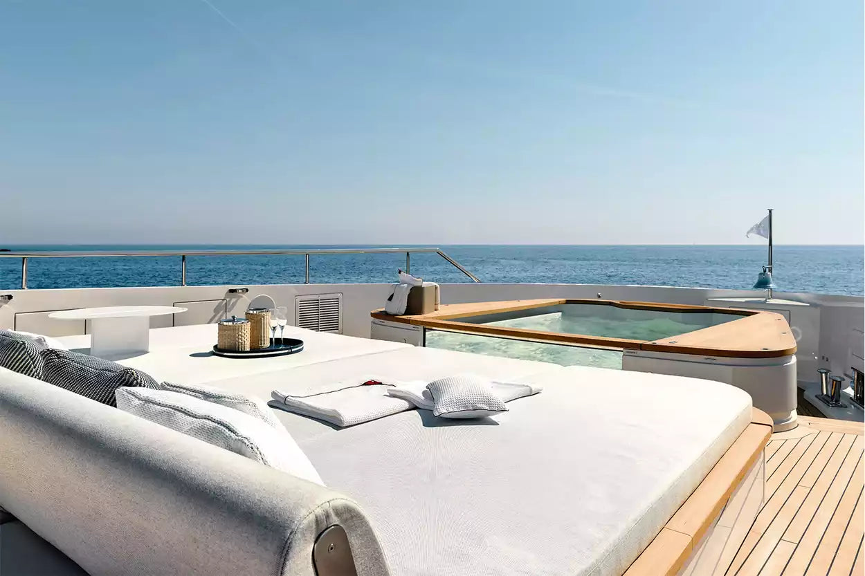 Luxury yacht outdoor lounge area with a large white daybed, matching pillows, a small built-in plunge pool, and an open view of the blue ocean under a clear sky.
