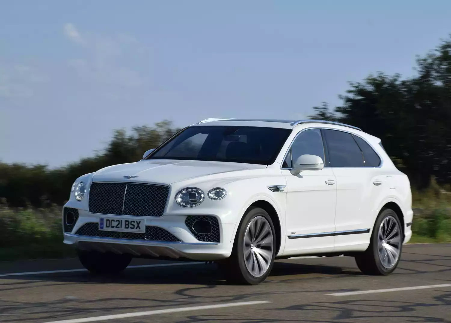 A white Bentley Bentayga SUV driving on a paved road with trees and blue sky in the background.