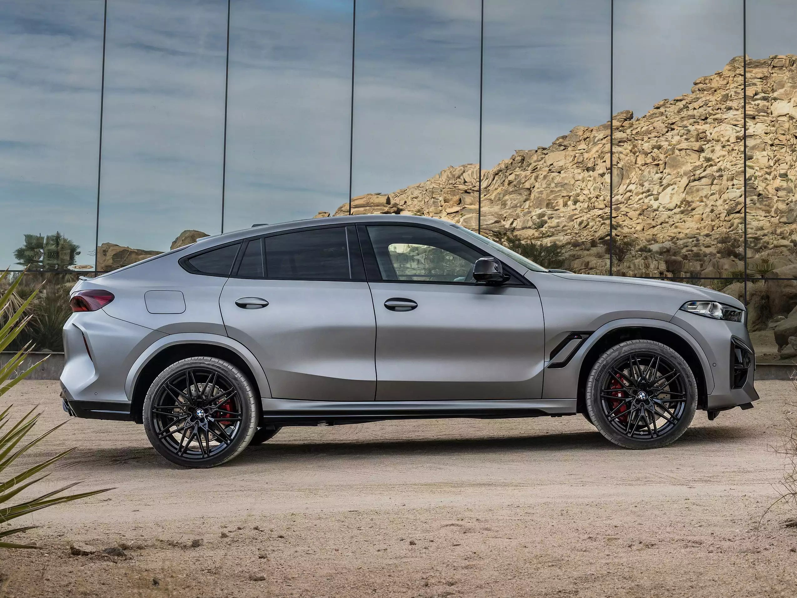A silver BMW X6 M is shown in a side profile view, parked outdoors in front of a modern glass wall with a rocky desert background.
