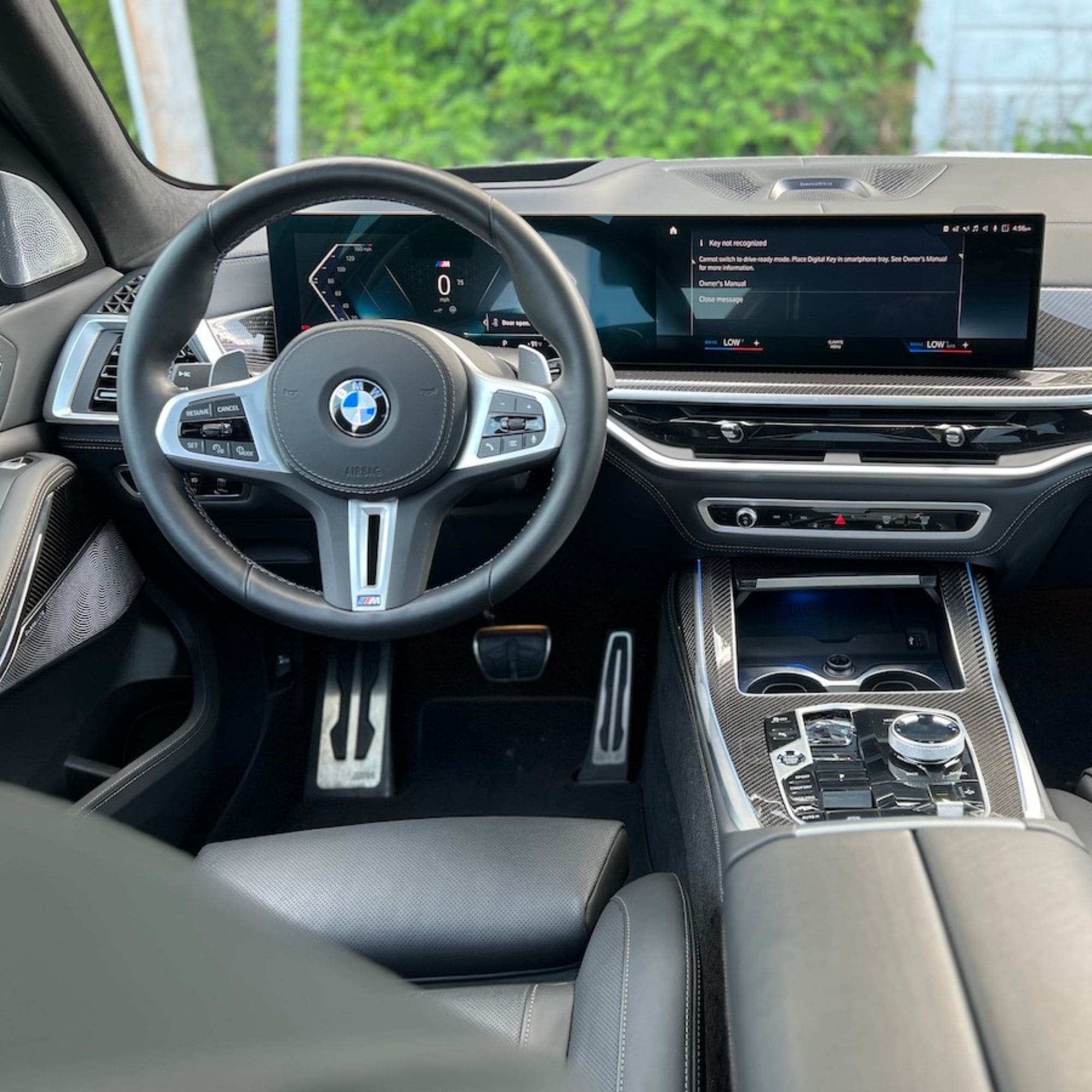 A luxury car interior with a black leather steering wheel featuring the BMW logo, a large digital dashboard display, and a center console with a rotary controller and carbon-fiber trim.