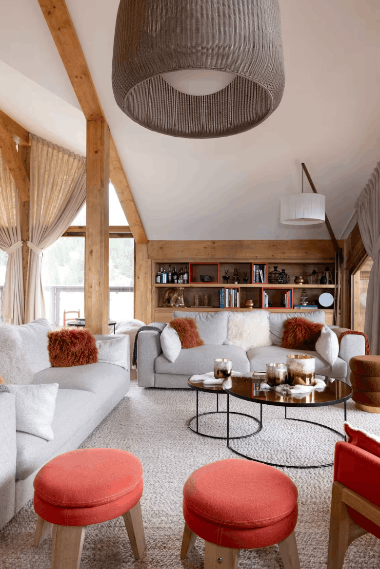 A bright, modern mountain-style living room with a white sofa, a red armchair, two orange-red ottomans, a round coffee table, and a large ribbed gray pendant light hanging from exposed wooden beams.