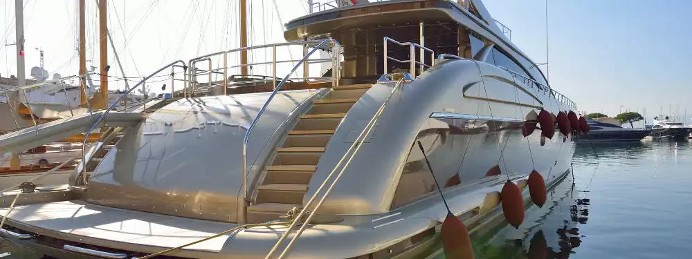 A close-up side view of a white luxury yacht docked in a marina, showing the rear staircase, metallic railings, and side fenders against a clear blue sky.