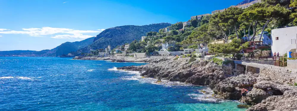 Coastal scene with blue ocean, rocky cliffs, and buildings on a hillside.