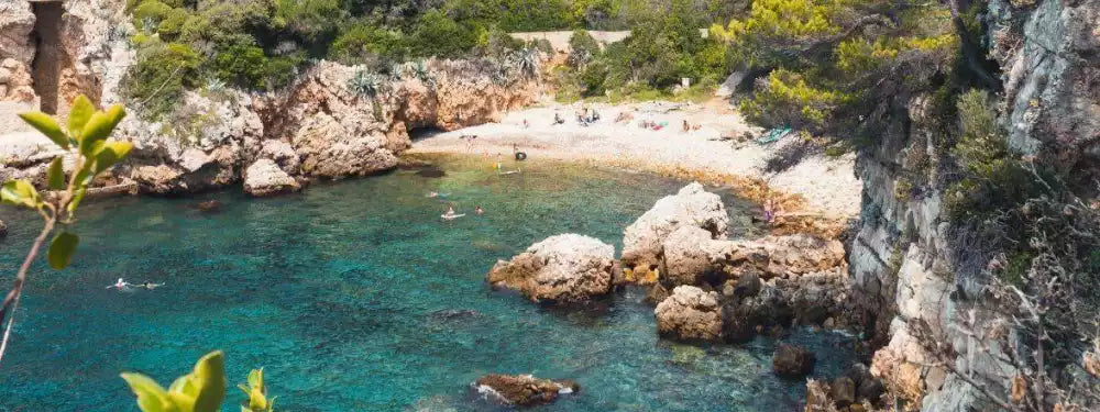A scenic coastal cove with turquoise water, rocky cliffs, a small sandy beach, and green vegetation under bright sunlight.