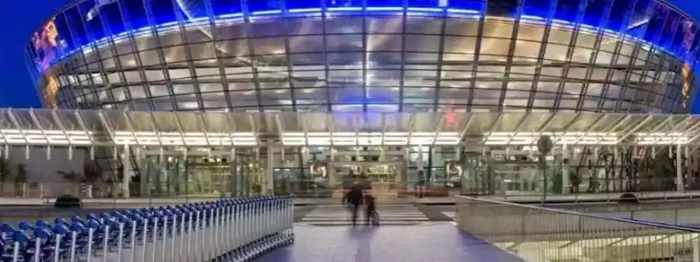 A low-angle night photograph of a modern, brightly lit stadium or arena with a curved glass facade, blue LED lighting, and a walkway in the foreground with a few blurred people.