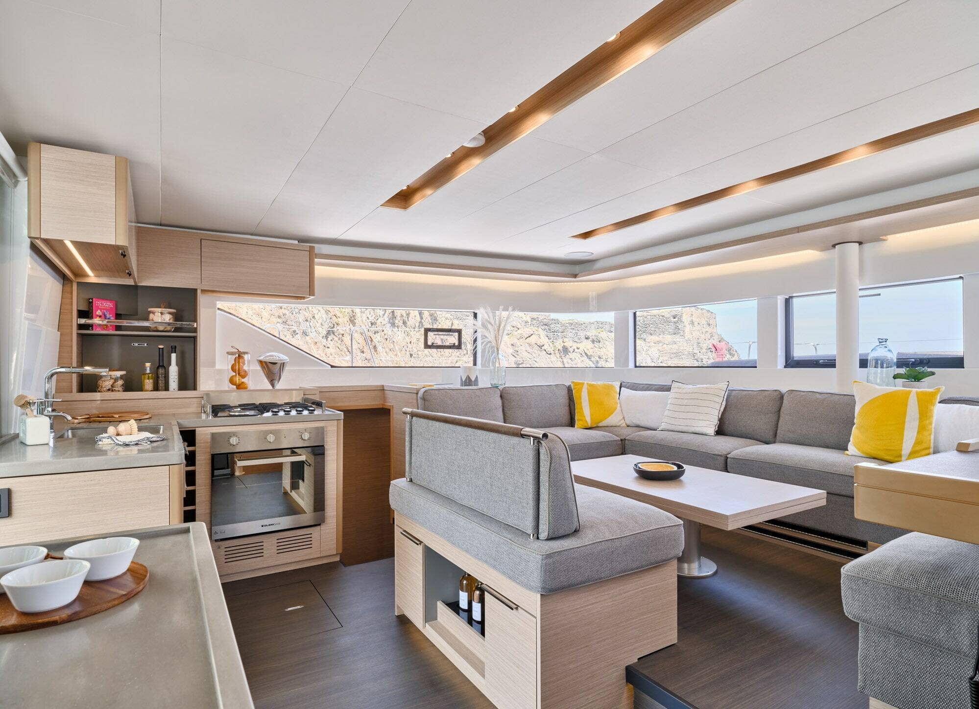 A modern yacht interior featuring a bright living area with a gray sectional sofa, white coffee table, and yellow accent pillows, alongside a compact galley kitchen with light wood cabinetry and stainless steel appliances.