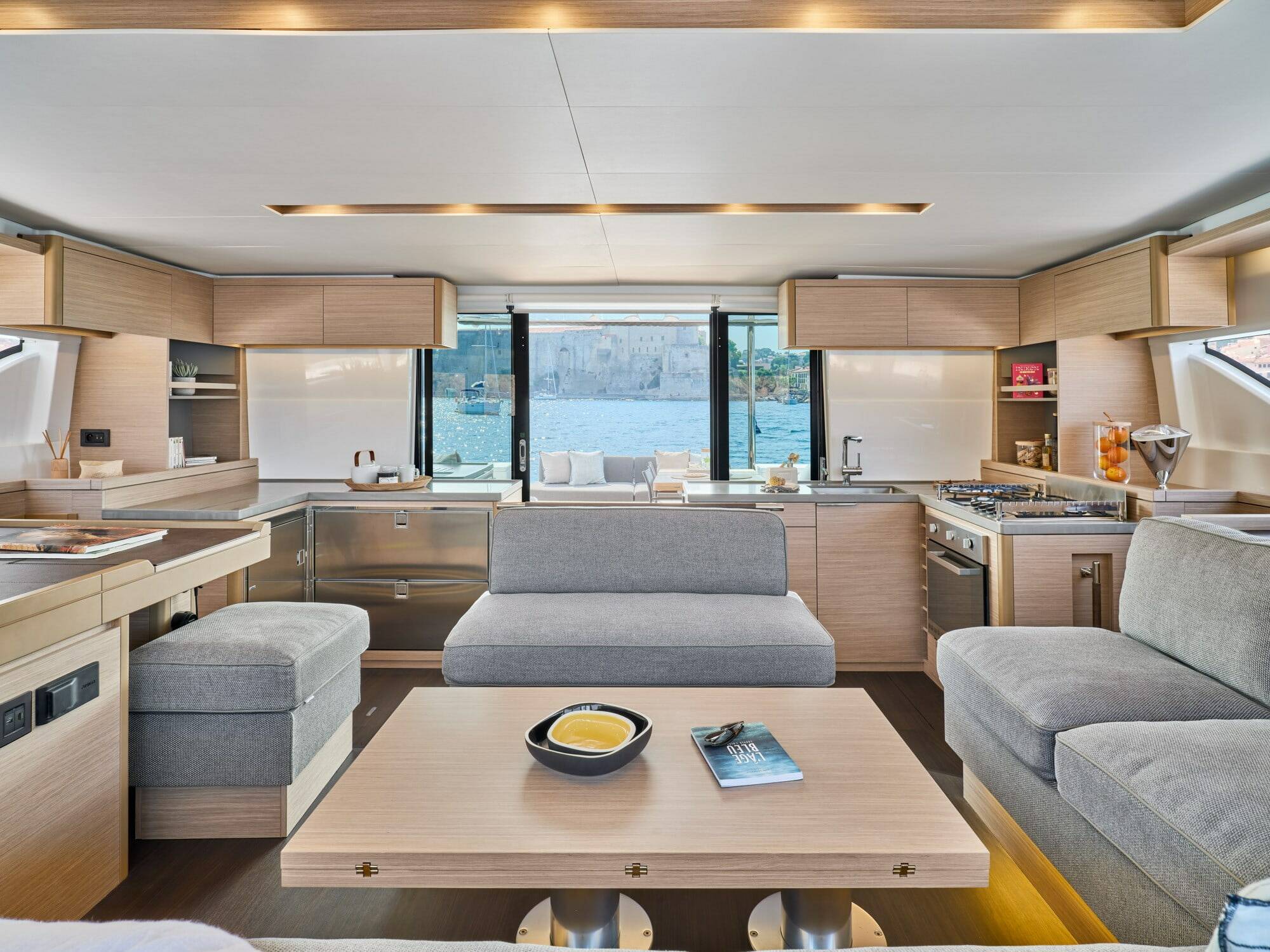 Modern yacht interior with a galley kitchen, seating area, and large windows overlooking water.