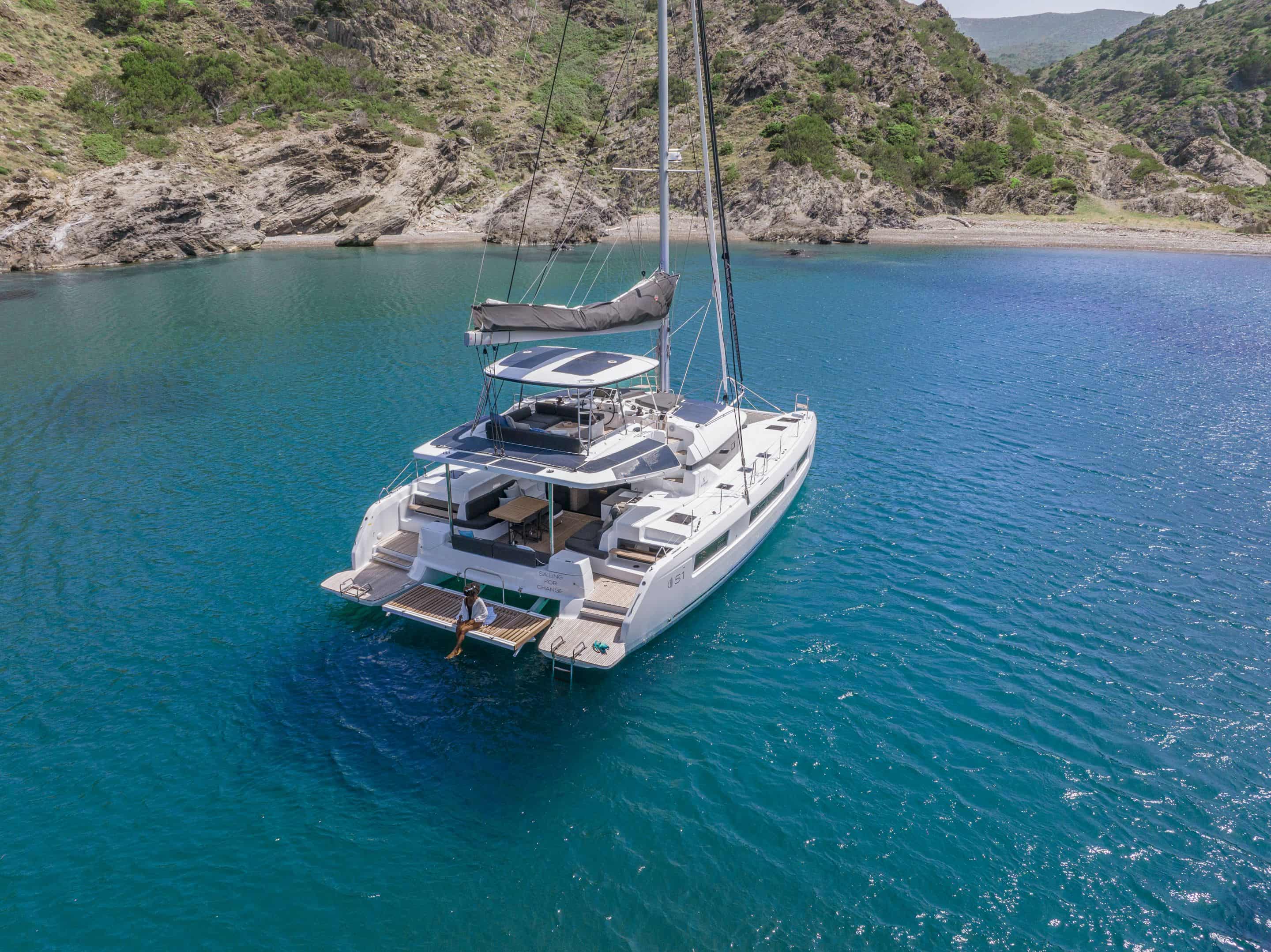 A white catamaran sailboat floating in clear turquoise water, with rocky green hills surrounding the bay. The boat has its sails down and a small platform extends from the rear, with a person visible standing on it.