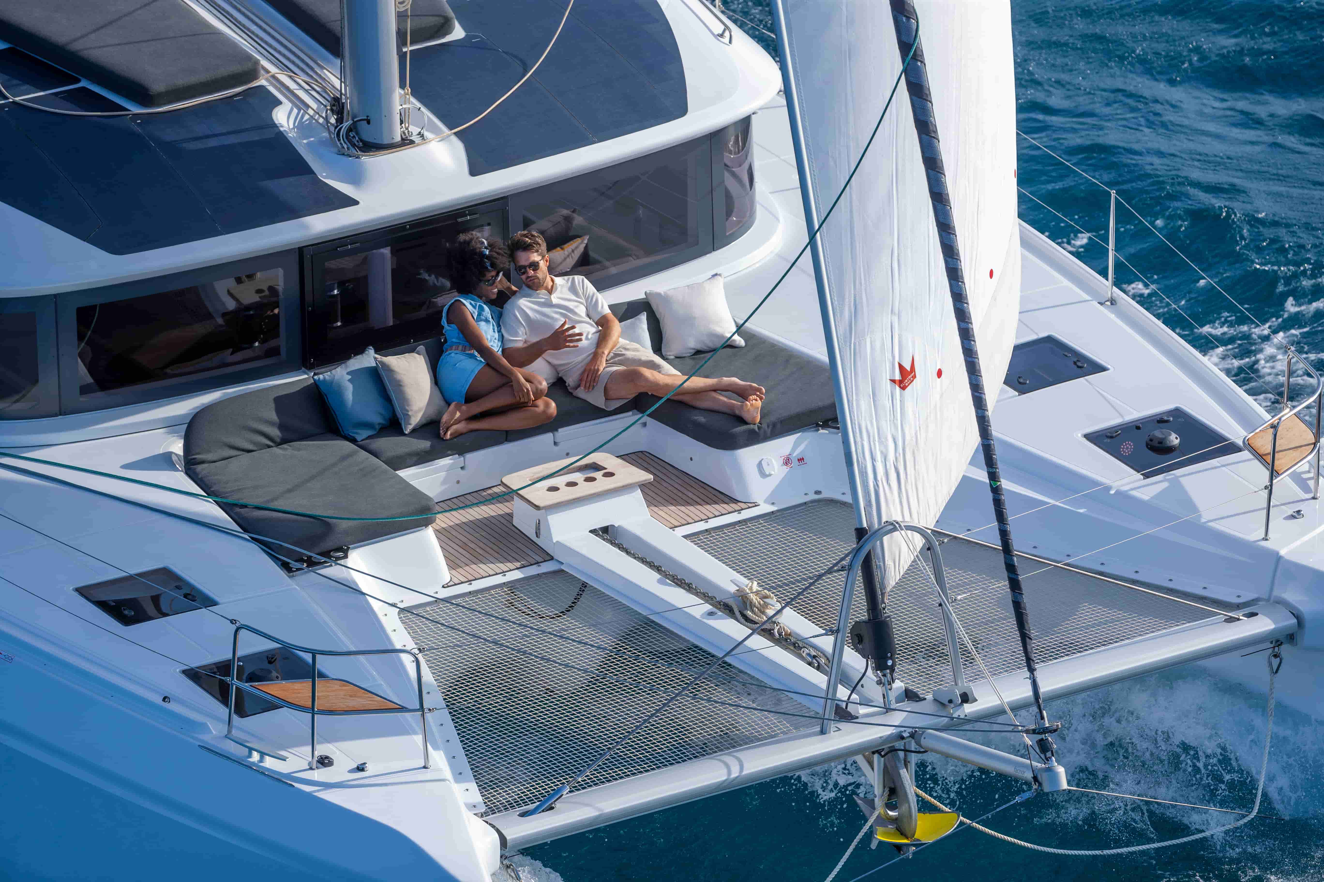 A couple relaxes on the deck cushions of a white catamaran yacht with a tall white mainsail, sailing on blue ocean water.