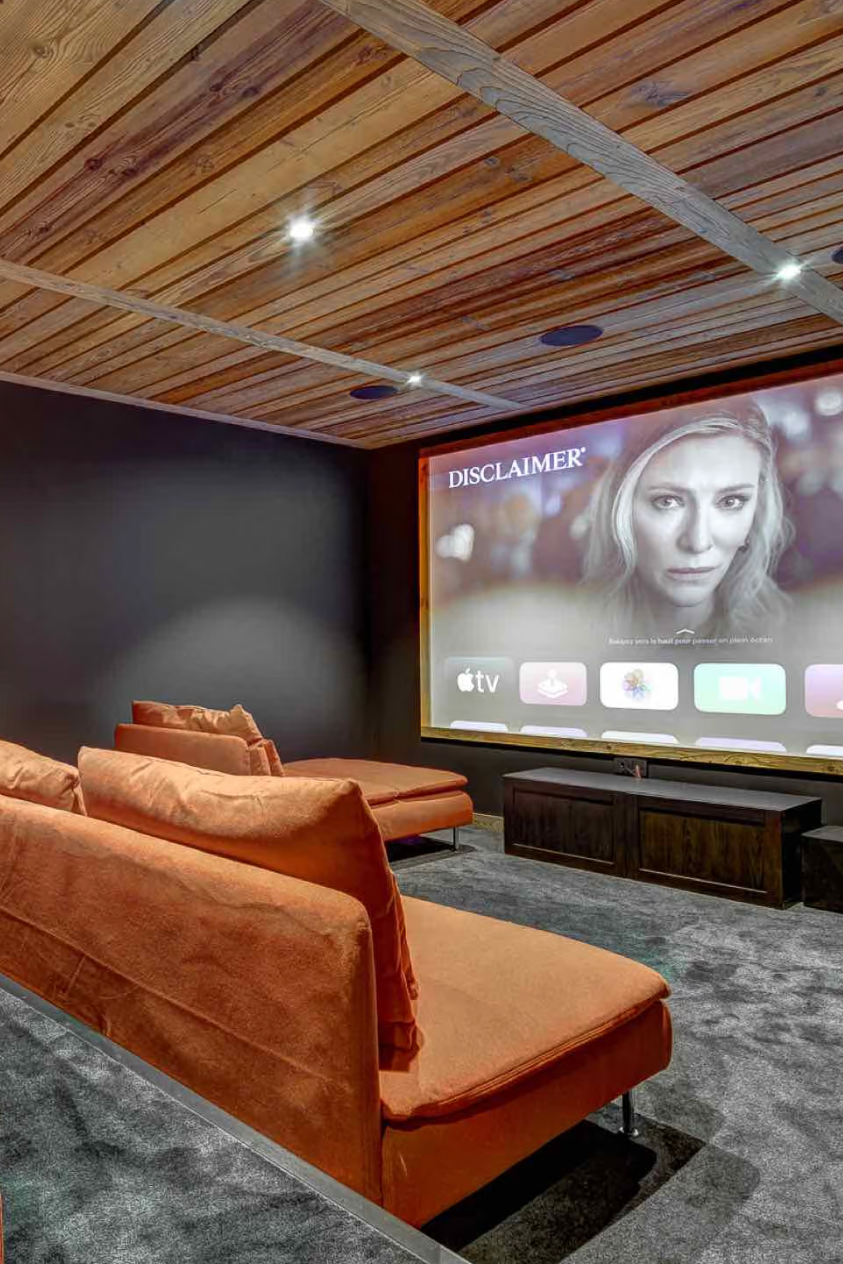 A home theater setup featuring plush orange sectional sofa seating, a dark gray carpet, and a large projection screen mounted on a dark wall under a wood-paneled ceiling.