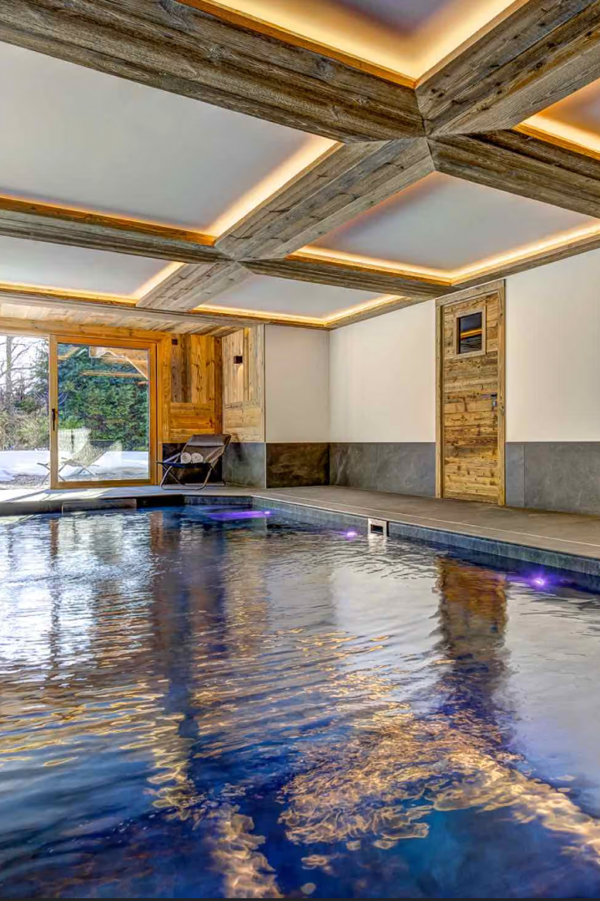 An indoor heated swimming pool with blue water and warm ambient lighting, surrounded by modern stone walls and a wooden ceiling with recessed lights.