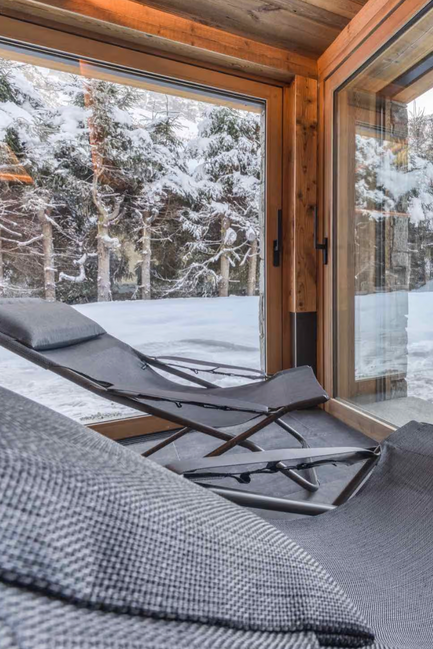Two dark gray outdoor lounge chairs positioned inside a wooden cabin with large windows overlooking a snowy outdoor area and winter trees.