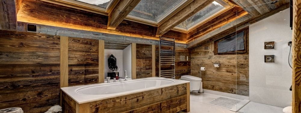 A luxury rustic bathroom featuring a large white bathtub set in a wooden cabin-style room with exposed wood paneling, a glass shower enclosure, and a skylight ceiling.