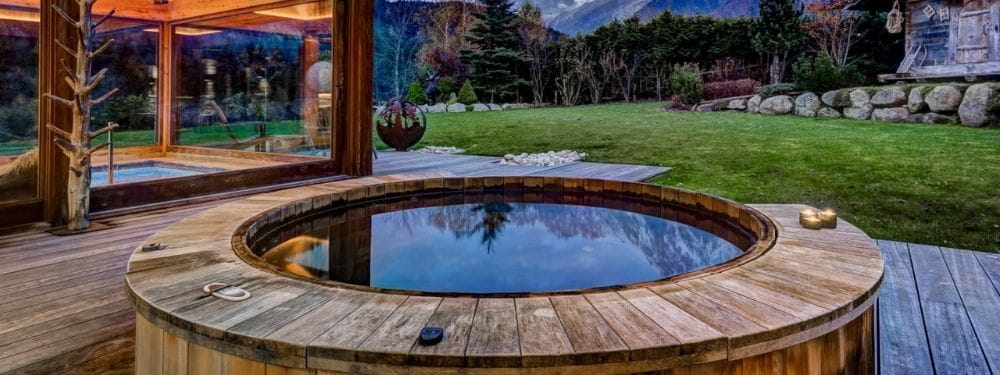 A round wooden hot spring or outdoor spa tub with still water, set on a wooden deck beside a glass-walled structure, with a manicured lawn and trees in the background.