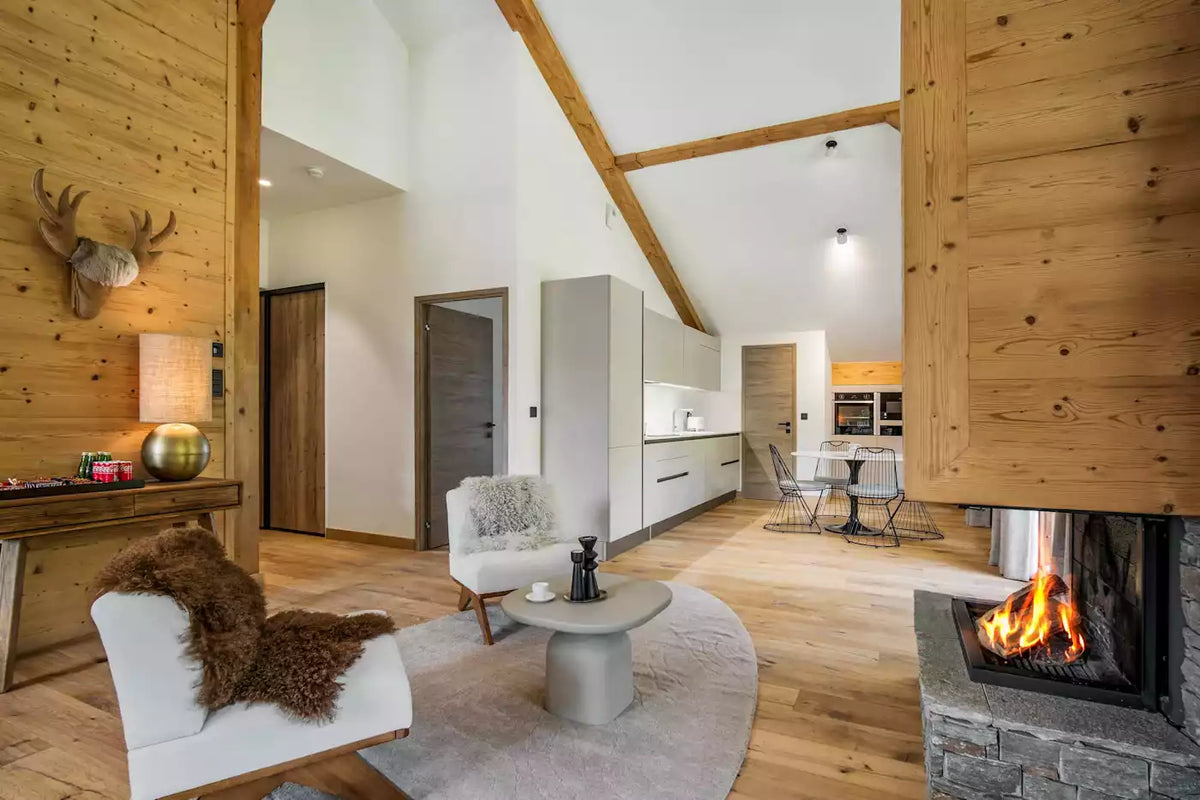 A modern open-plan chalet interior with light wood walls and ceiling beams, white chairs, a round coffee table, a visible fireplace with flames, and a kitchen area in the background.