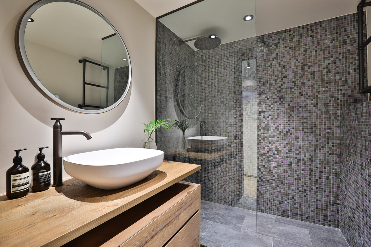 Modern bathroom vanity with a white vessel sink, dark wood countertop, round wall mirror, and a walk-in shower area covered in gray mosaic tile.