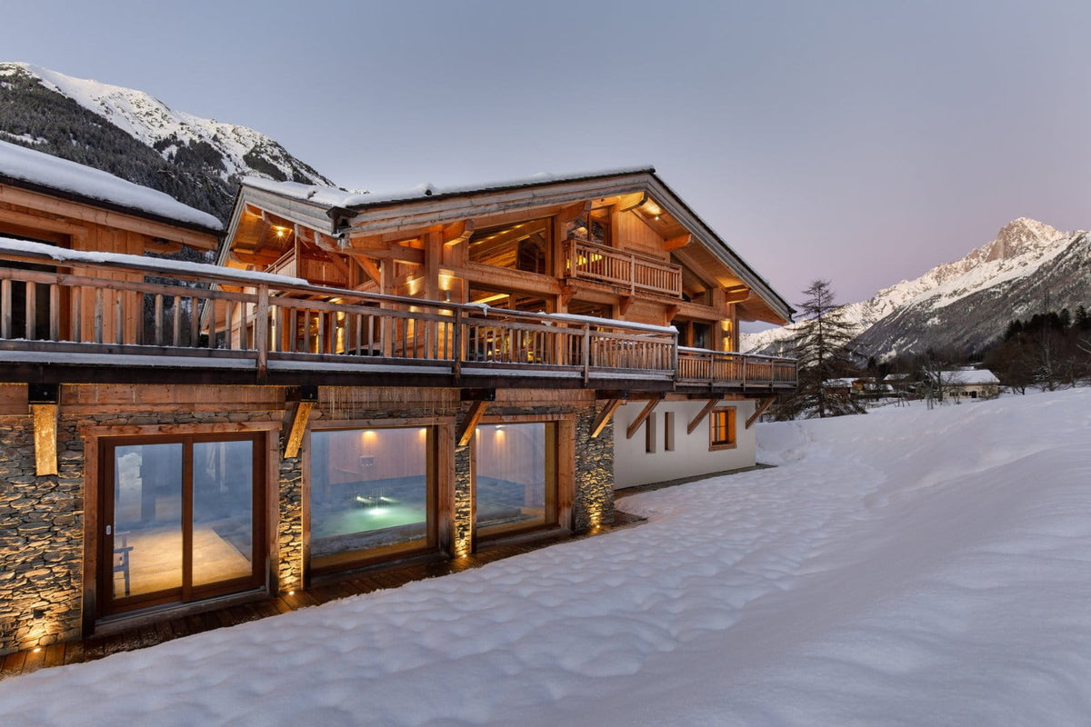A luxury alpine chalet with warm glowing windows and wooden balconies, nestled in a snowy mountain landscape at dusk.