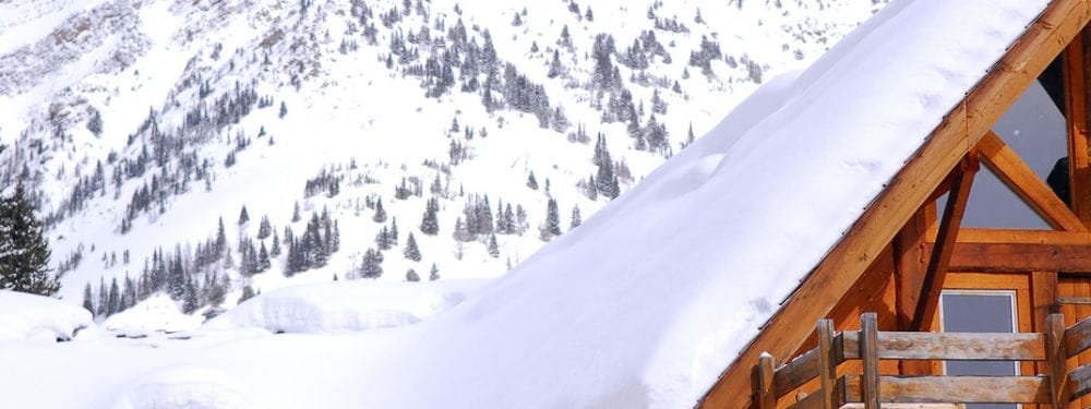 A snowy mountain landscape with a wooden chalet in the foreground, featuring a snow-covered roof and railing against a backdrop of snow-covered peaks and evergreen trees.