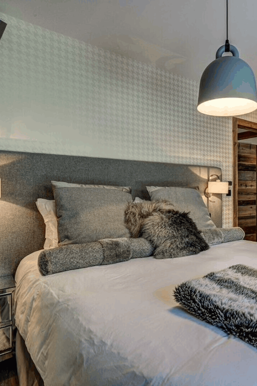 A modern bedroom featuring a bed dressed in white bedding with gray and black textured pillows and throws, set against a gray upholstered headboard. The room includes a rustic wood-paneled wardrobe, a textured light gray wall, bedside tables with lamps, and a hanging pendant light.