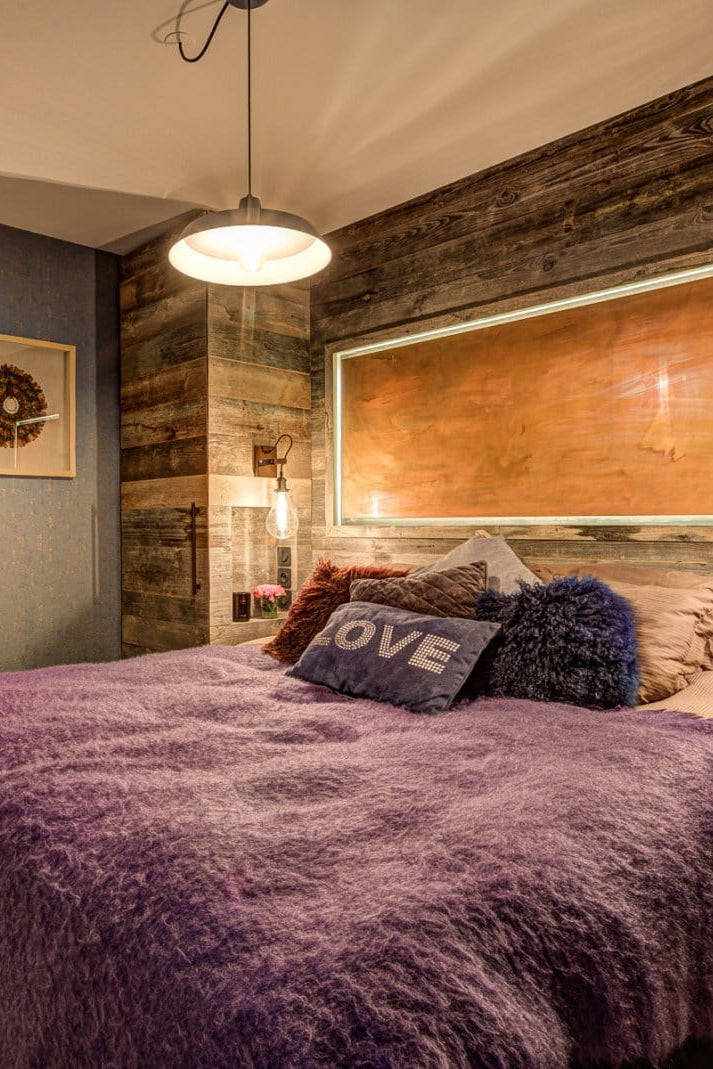A rustic bedroom featuring a bed with a textured purple shag blanket and pillows, set against a wall made of dark wood planks with an integrated light. A blue accent wall and a wooden door are visible on the left.