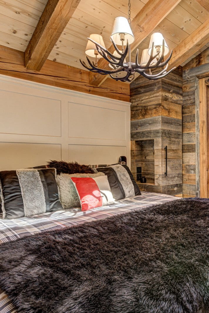 A rustic bedroom interior with a large bed dressed in plaid bedding and a dark faux-fur throw blanket, set in a cabin-style room with wood-paneled walls, a chandelier, and a glass shower enclosure in the background.