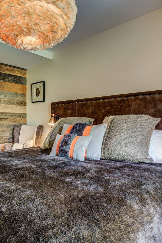 A rustic-style bedroom featuring a large bed with a textured brown faux-fur duvet, a dark brown leather headboard, and a mix of gray, blue, and orange accent pillows. The room has warm beige walls, a large woven pendant light, and a geometric orange and white rug.