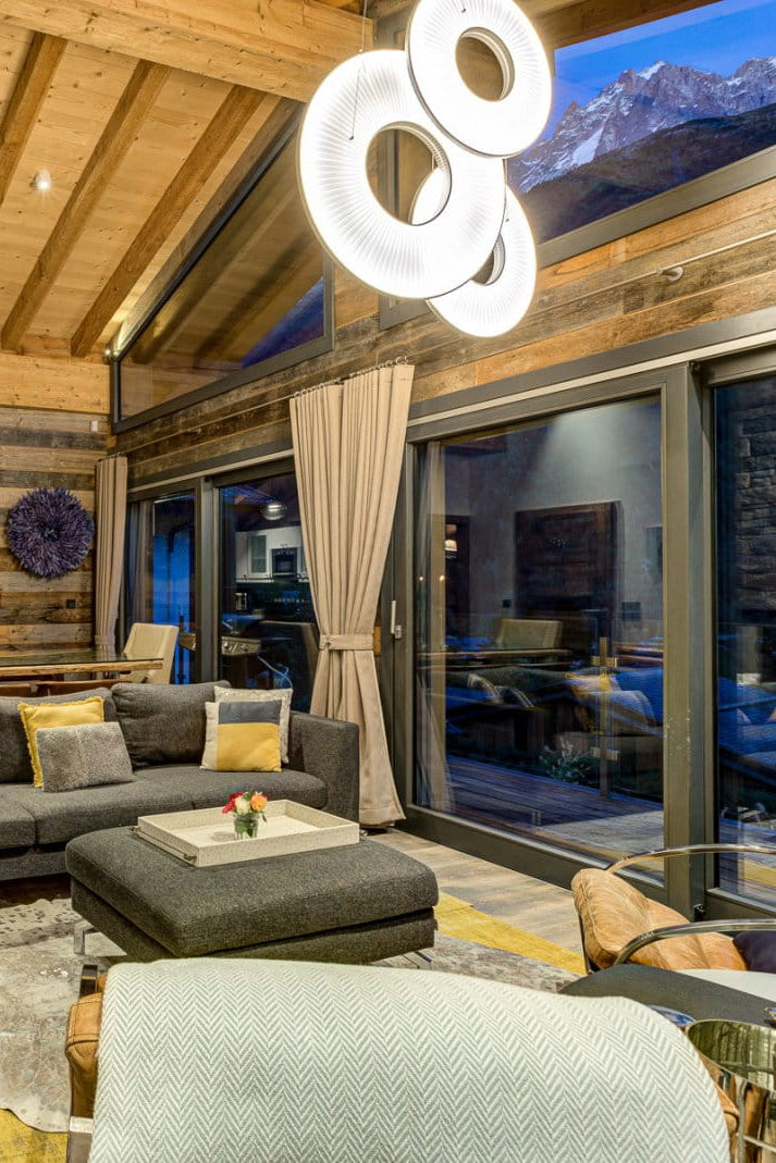 A luxury modern chalet living room featuring a dark gray sectional sofa with yellow accent pillows, a stone fireplace, a high wood-beamed ceiling, and large windows looking out onto a mountain view.