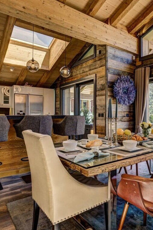 A luxury alpine chalet dining room featuring a long wooden dining table set with white dishes, surrounded by brown leather and cream upholstered chairs, with a kitchen and stunning mountain views through large windows.