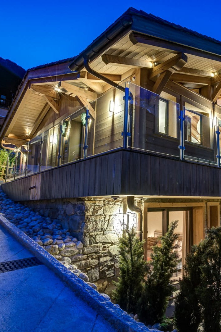 A luxury chalet-style mountain lodge at dusk, featuring warm wood and stone exterior walls, illuminated windows, a large wooden deck with outdoor seating, and snow-covered slopes in the background under a deep blue twilight sky.