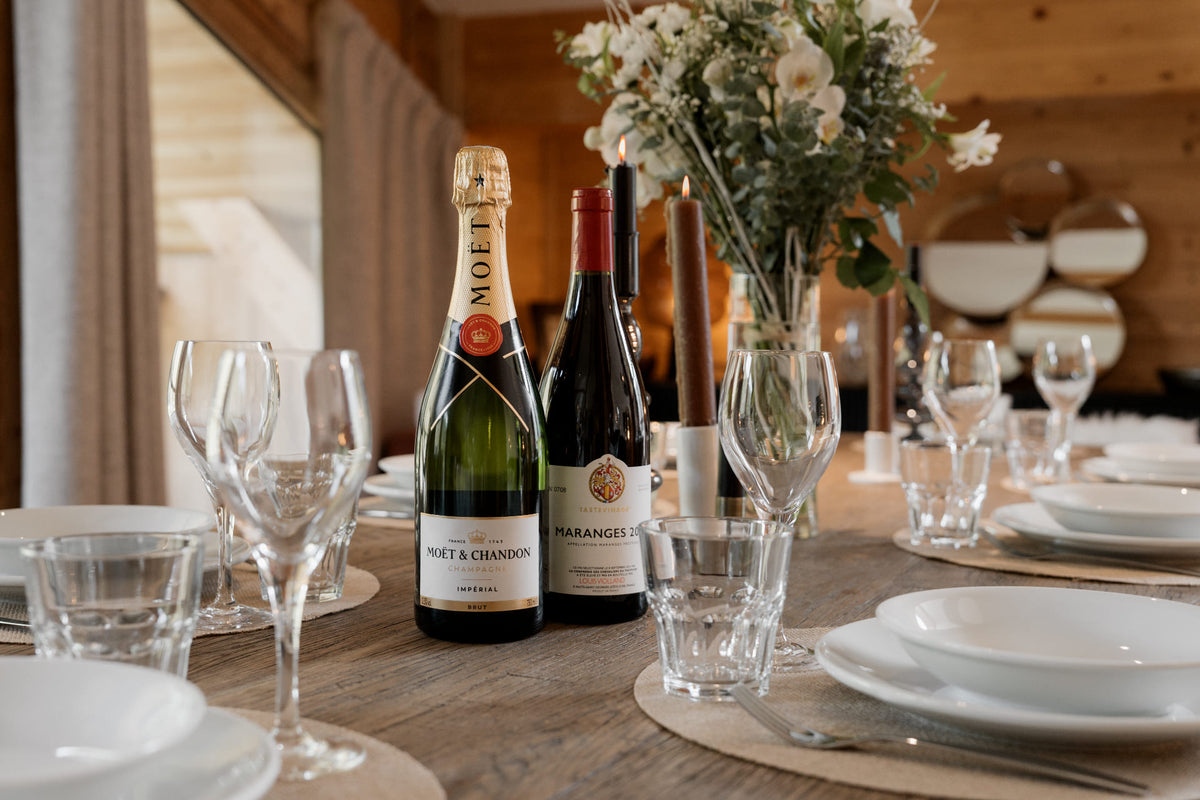 A Moët & Chandon Brut Champagne bottle and a dark red wine bottle set on a rustic wooden dining table surrounded by white plates, clear wine glasses, and a floral centerpiece.