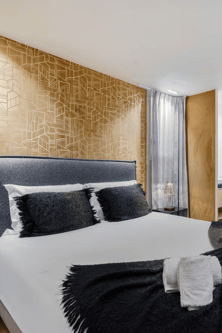 A modern bedroom featuring a bed dressed in white linens with dark gray pillows and a dark gray throw blanket, set against a geometrically patterned gold accent wall and a large light wood wardrobe.