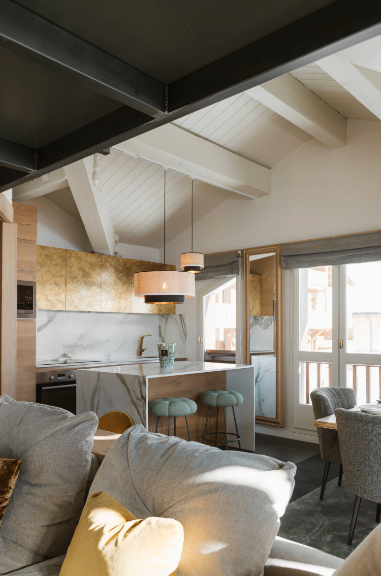 A modern open-plan living and kitchen area with a gray sofa in the foreground, pendant lights, marble countertops, and a high-ceilinged room with exposed beams.