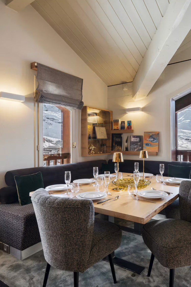 A modern ski chalet dining room featuring a wooden dining table set with white plates and wine glasses, surrounded by textured gray upholstered chairs. A dark gray L-shaped banquette sits along the back wall.