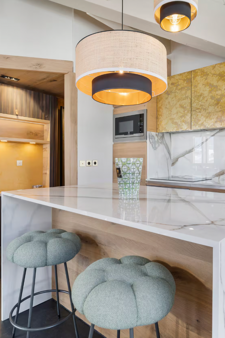 A modern kitchen island with a white marble countertop and base, paired with two round tufted gray bar stools. The background features a kitchen with gold-leaf patterned cabinets, a marble backsplash, and warm wood cabinetry.