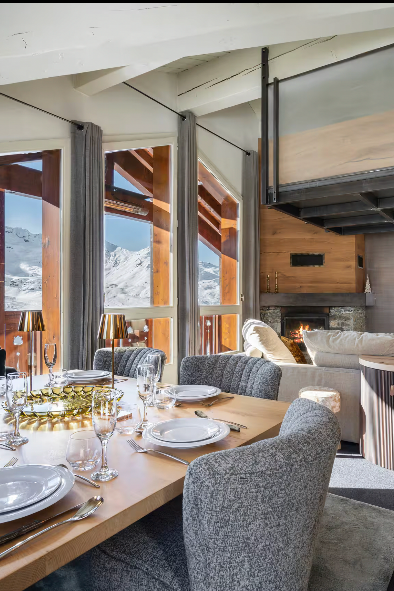 A luxury modern mountain chalet interior featuring a dining area with a wooden table set for six, gray upholstered chairs, large windows overlooking snowy mountains, a loft bed with dark metal railings, a curved wood bar, and a yellow accent chair.