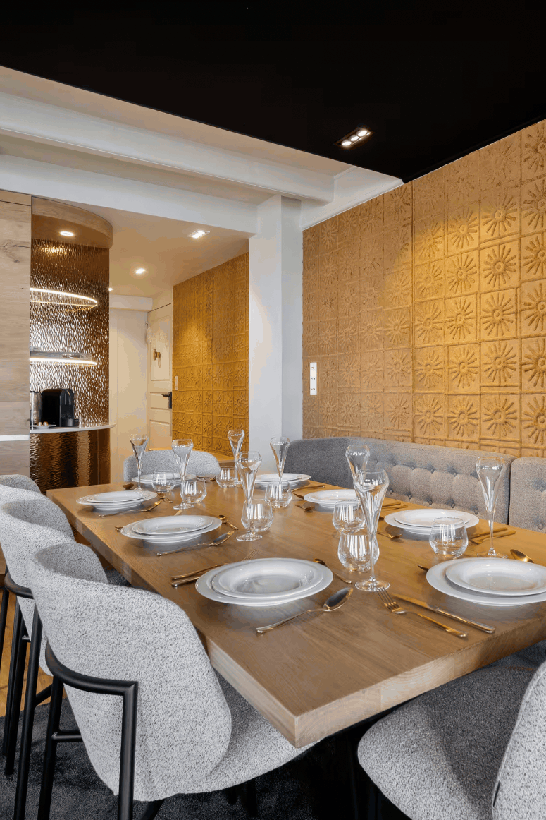 A modern dining room interior featuring a rectangular wooden table set with white plates and glassware, surrounded by gray upholstered chairs. A tufted gray banquette runs along one side, and the wall behind it is covered in warm gold-toned decorative panels.