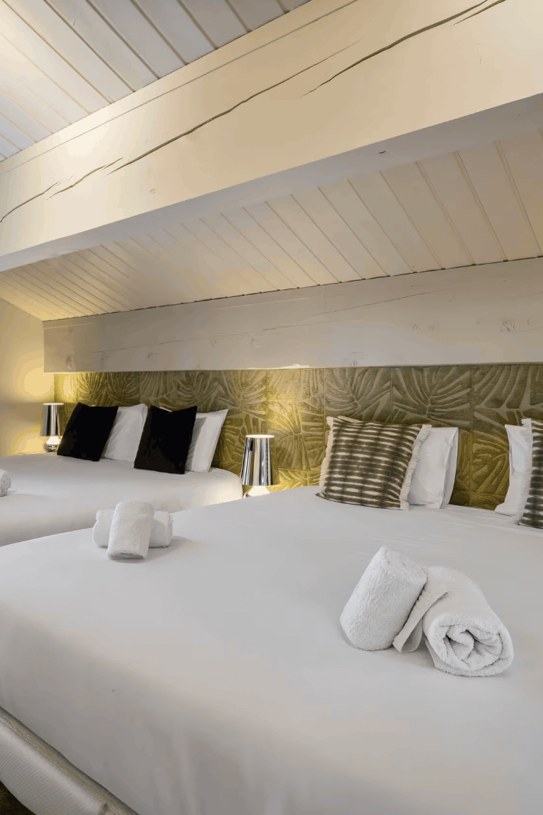 A luxury attic bedroom featuring two neatly made beds with white linens, decorative striped pillows, and folded towels, set against a green patterned headboard with warm ambient lighting.