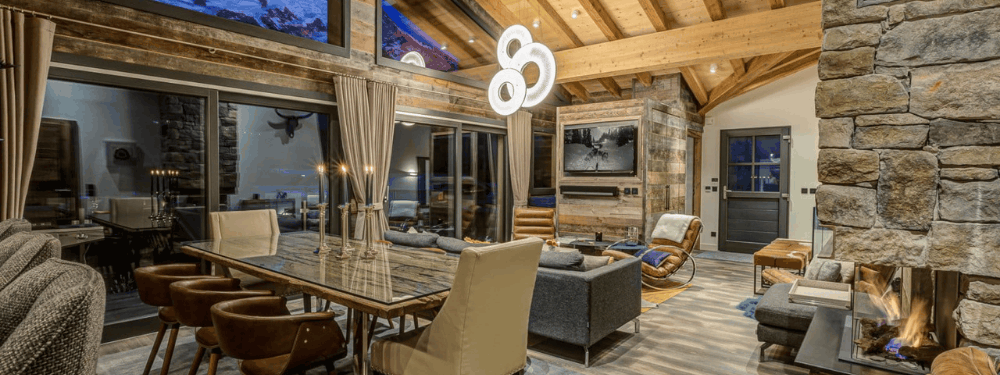 A luxury modern chalet interior featuring a dining area with a glass-top table and beige chairs, a stone fireplace with a fire, a gray sofa, and a stone wall with a TV mounted on it.