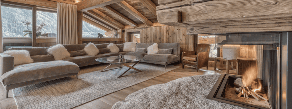 A luxurious alpine chalet living room featuring a large gray sectional sofa with fluffy white throw pillows, a round glass-top coffee table, a roaring fireplace with a dark metal surround, and a cozy wood-paneled interior with a mountain view.