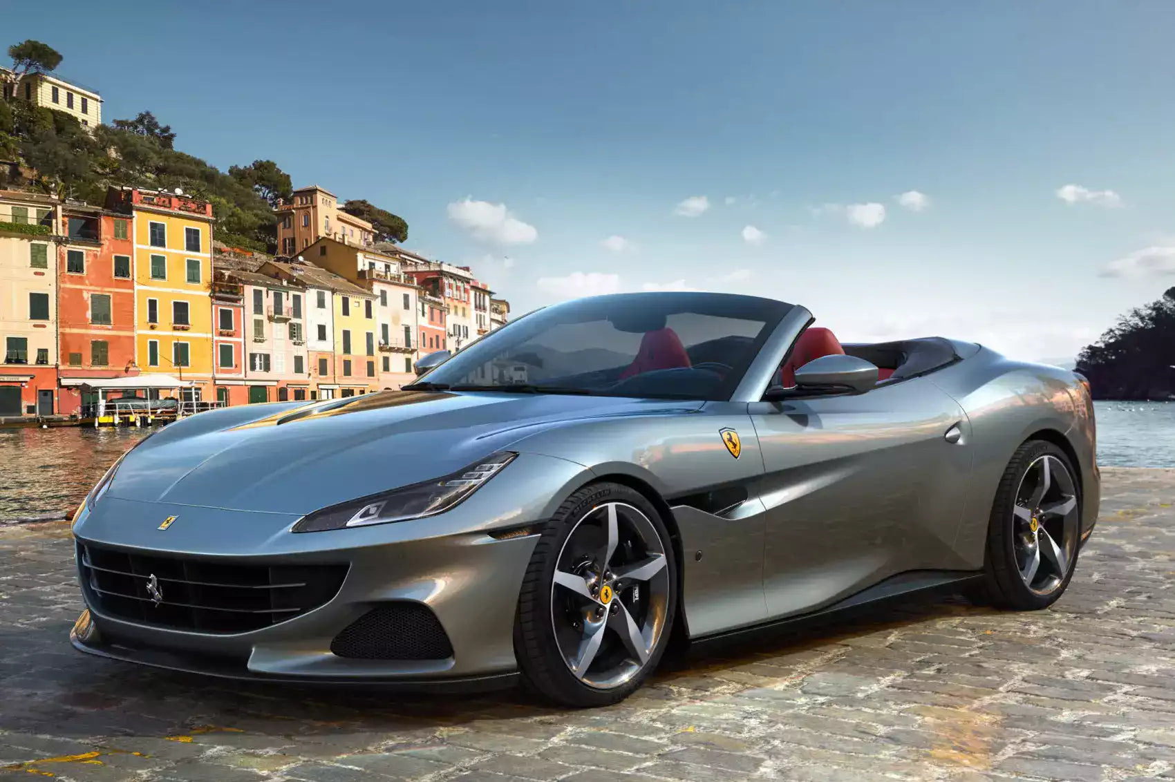 A silver Ferrari Portofoglio convertible sports car parked outdoors on a stone surface with a colorful coastal town and blue sky in the background.
