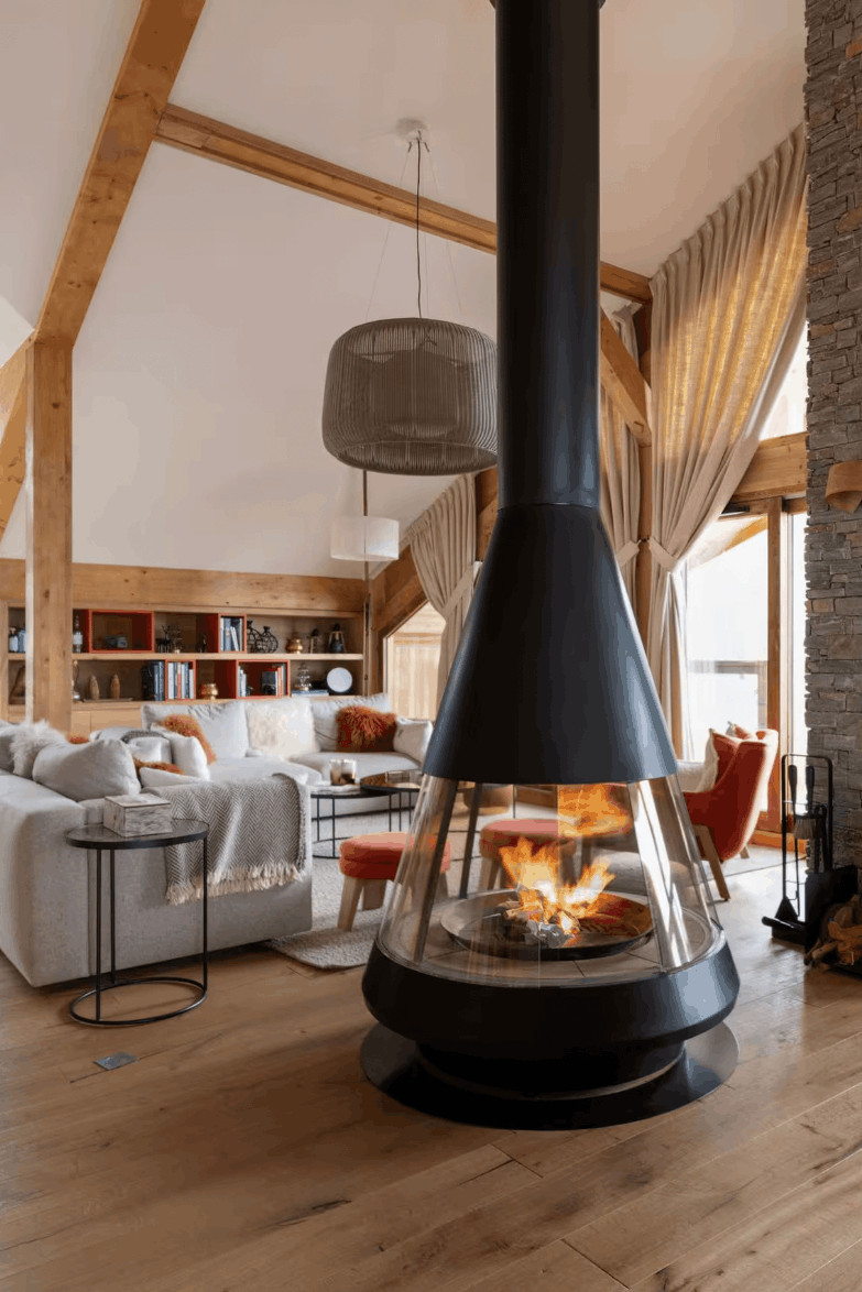 A modern black conical wood-burning stove with a glass front and visible fire, set in a rustic chalet-style living room with light wood floors, stone walls, and a fireplace mantel.