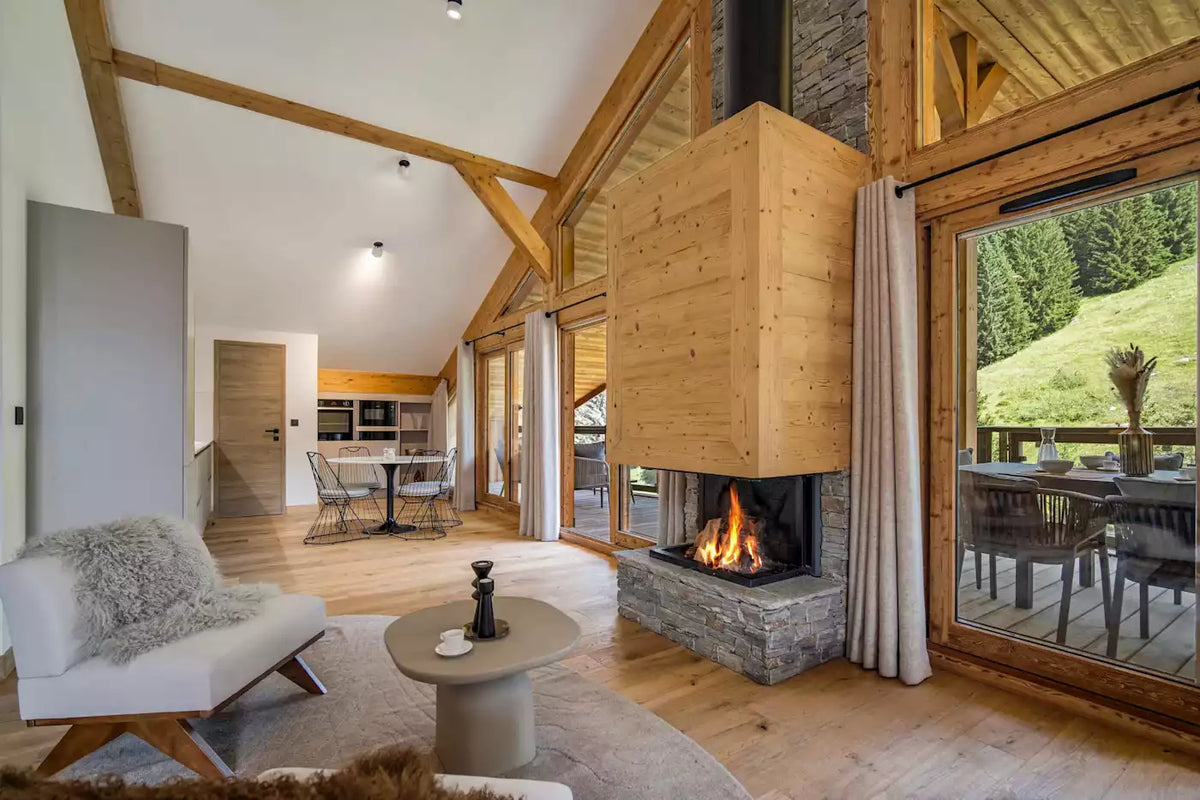 A modern luxury chalet interior featuring a large wooden cube fireplace with a stone base, high wood-beam ceilings, a white armchair with a gray shag pillow, and large windows looking out to a green mountain landscape.