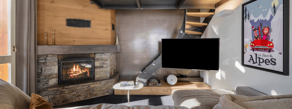 A cozy modern living room interior featuring a stone fireplace with a wood-burning fire, a mounted flat-screen TV, a staircase, and a framed 'Les Alpes' poster on the wall.
