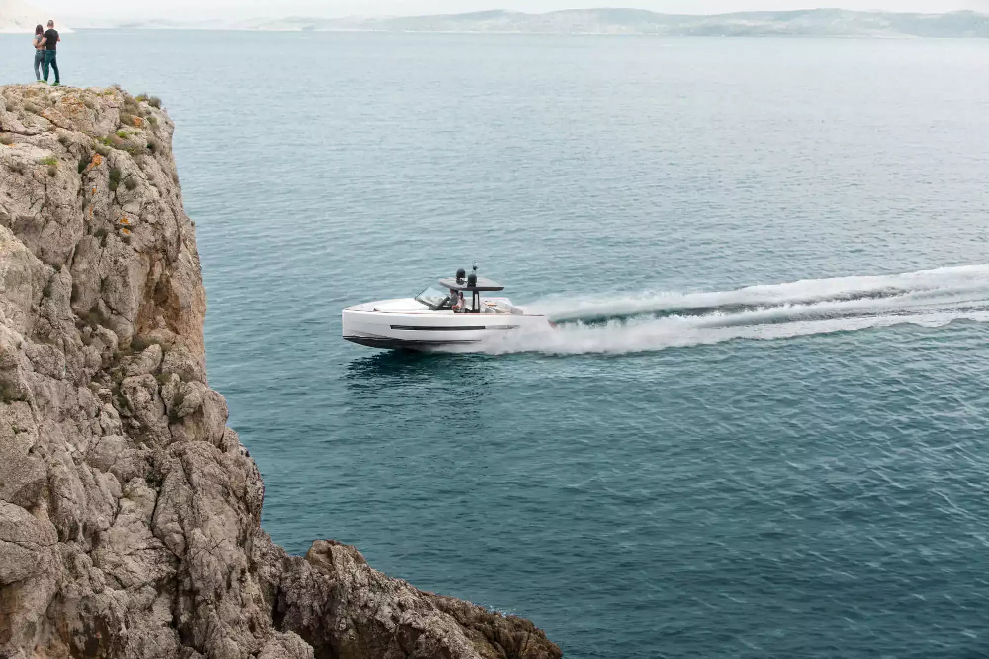 Speedboat on the water near a rocky cliff with people standing on the edge.