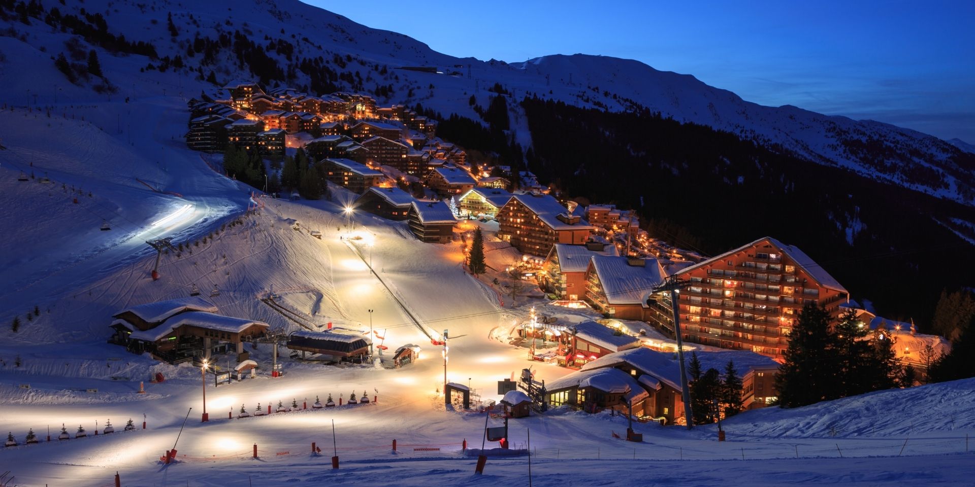 Snow-covered mountain village at night with illuminated buildings and ski slopes.