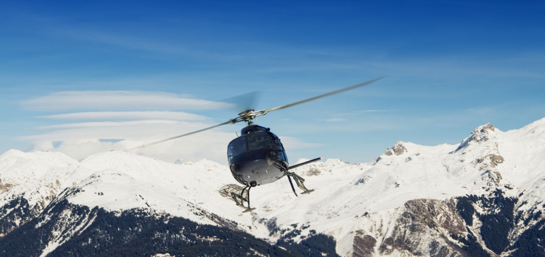 Helicopter flying over snow-covered mountains with a clear blue sky.