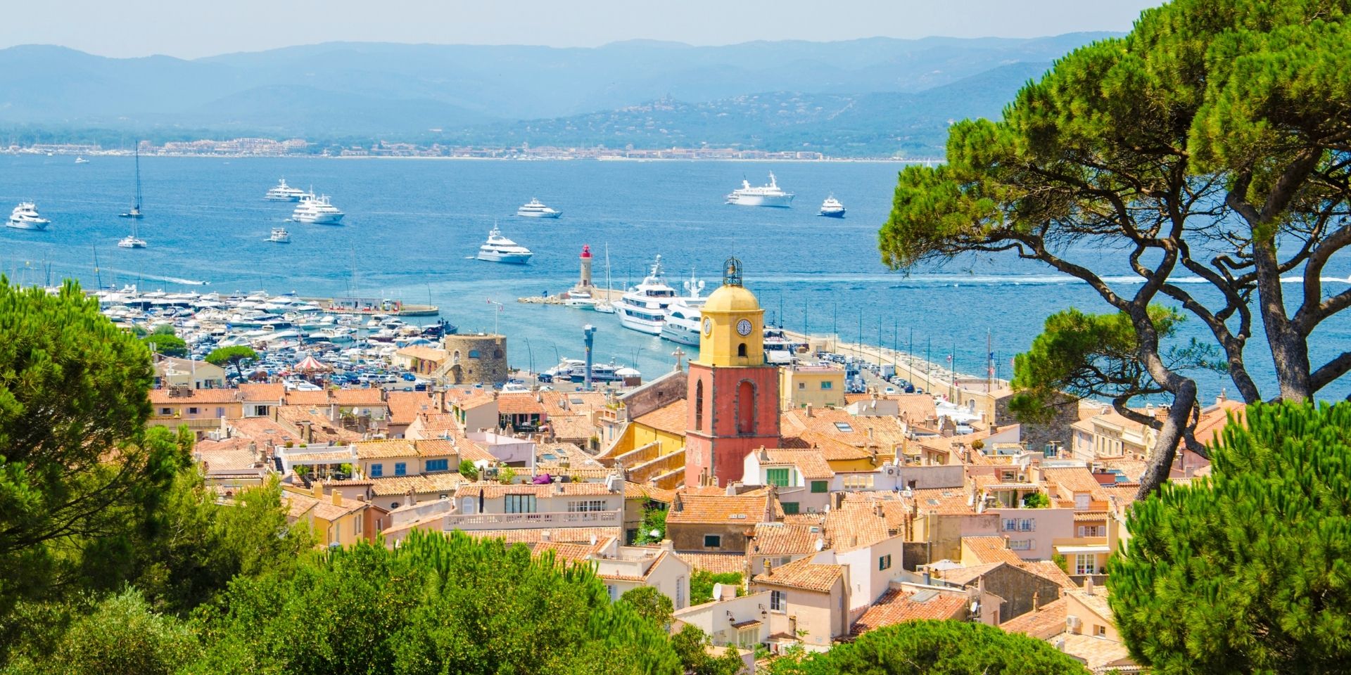 Village by a marina with boats and a lighthouse, surrounded by trees.