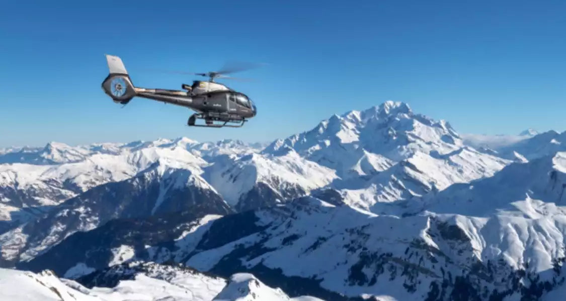 Helicopter flying over snow-covered mountains under a clear blue sky
