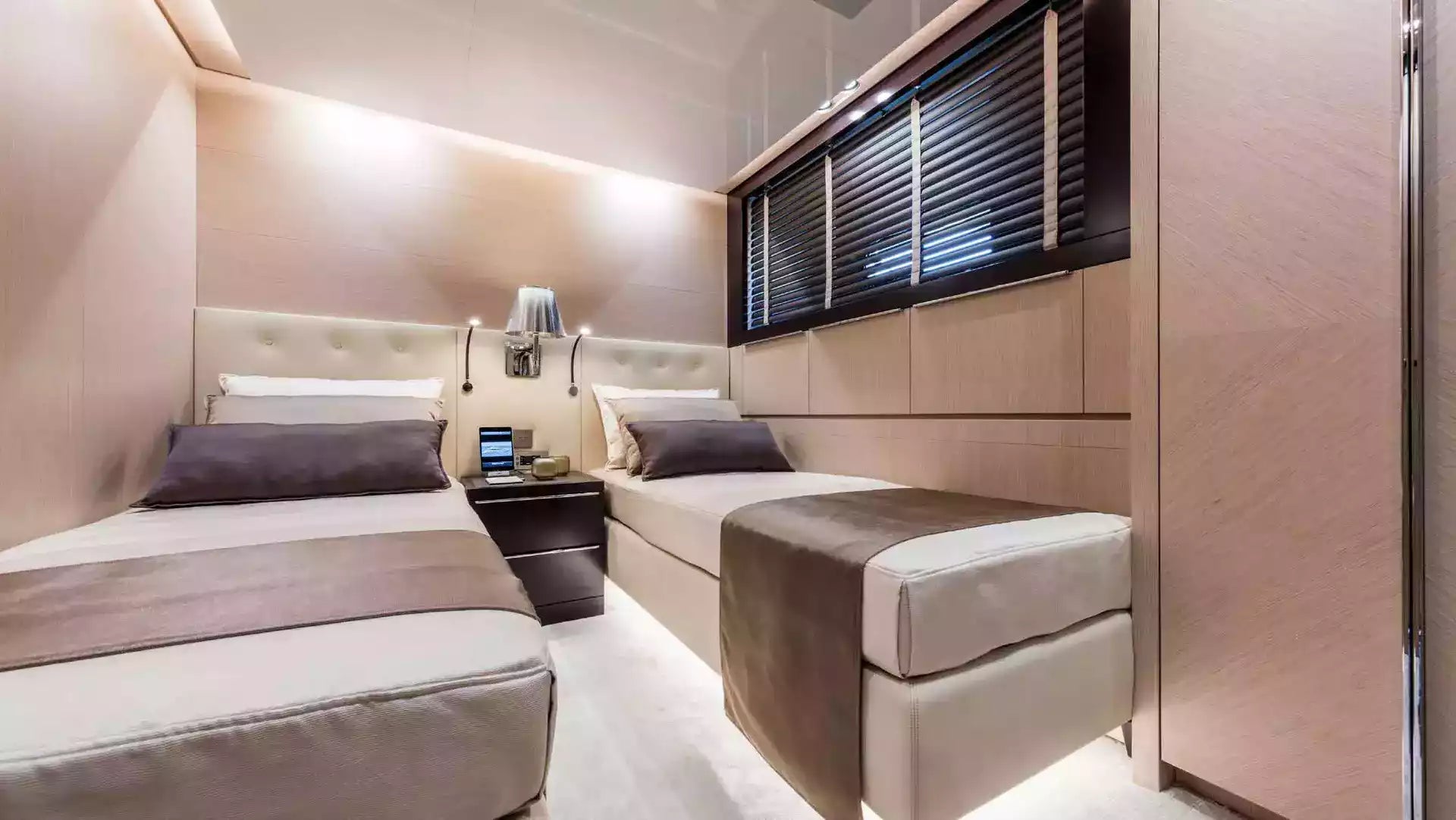 A luxury yacht or cabin interior featuring two neatly made beds with beige and brown bedding, a matching bench, a dark wood nightstand, and a large window with black blinds.